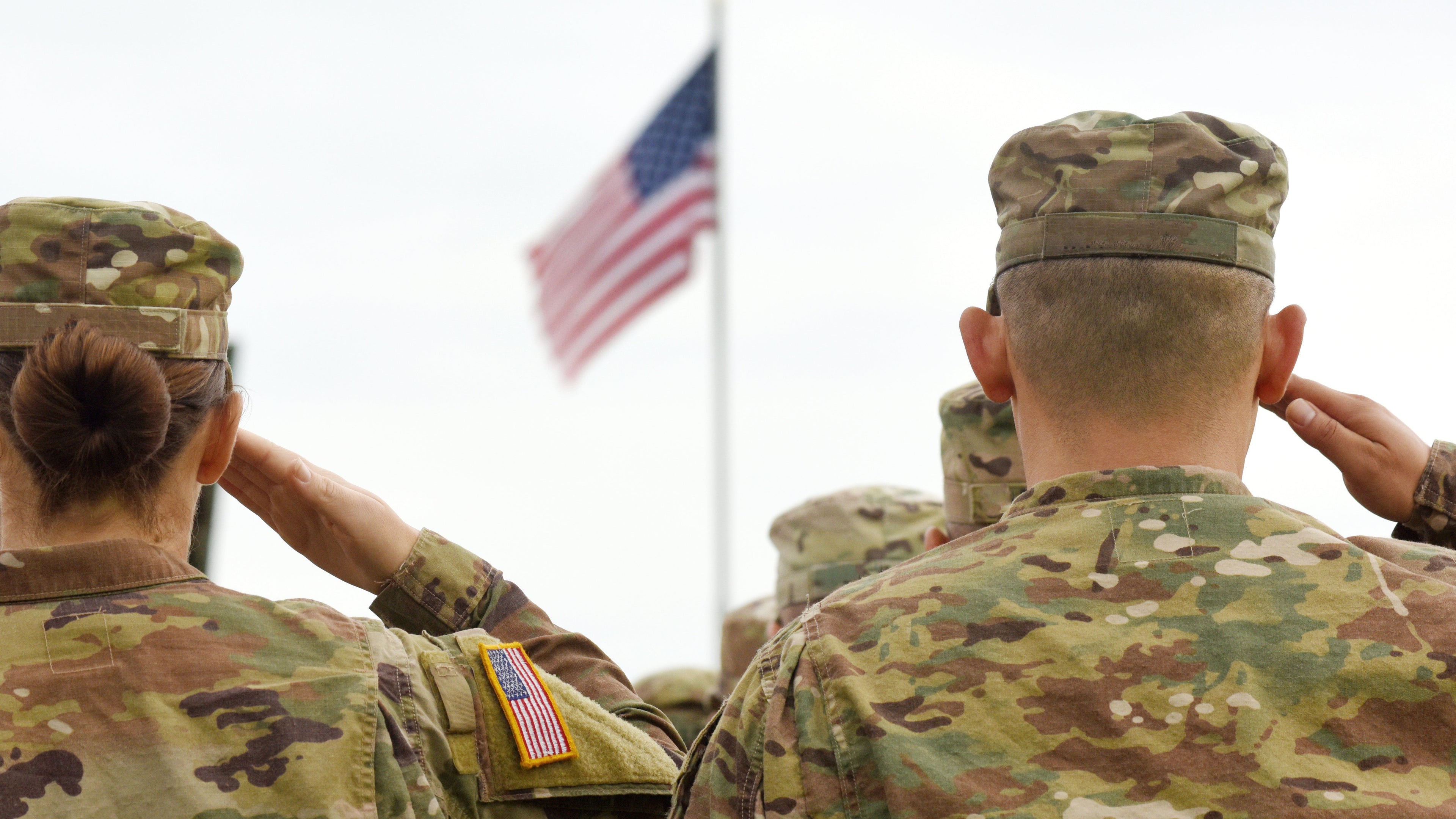 Soldiers in camouflage uniforms saluting with an American flag in the background